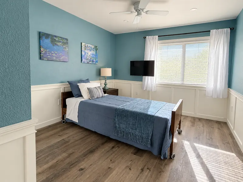A sunlit renovated resident room with soft blue walls, white wainscoting, wood floors and a TV
